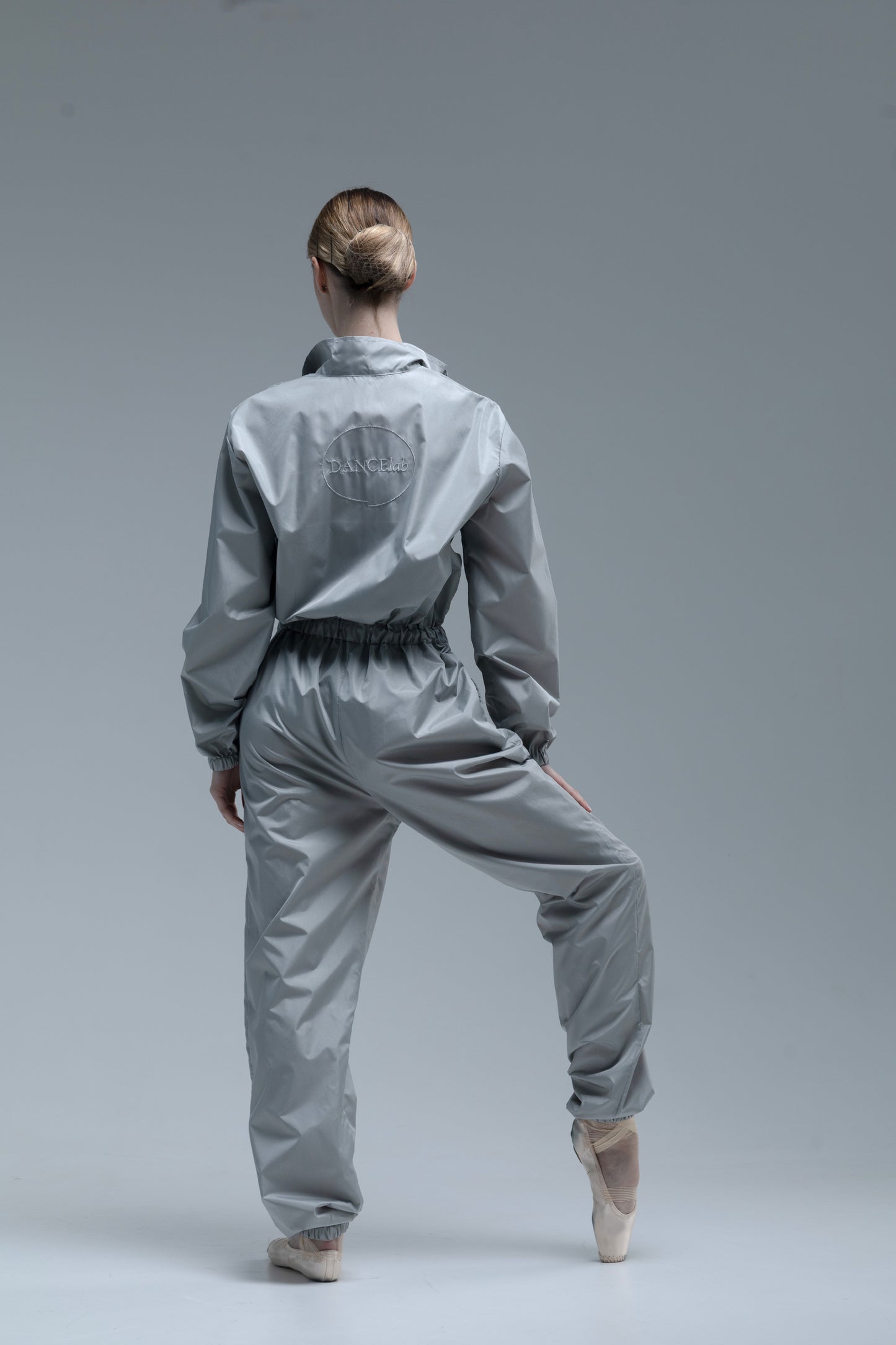 A ballerina model from behind wearing a shiny light gray dancewear set consisting of a cropped long-sleeve top and matching pants, posed on a seamless gray studio background.