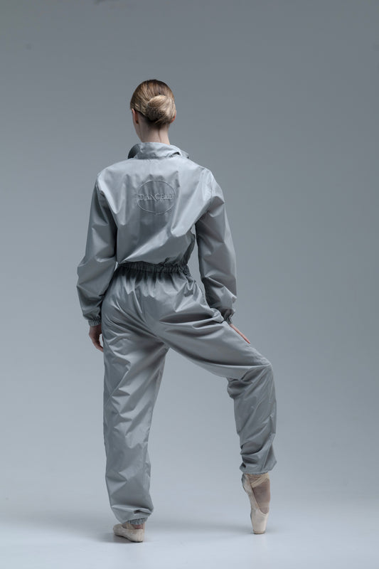 A ballerina model from behind wearing a shiny light gray dancewear set consisting of a cropped long-sleeve top and matching pants, posed on a seamless gray studio background.