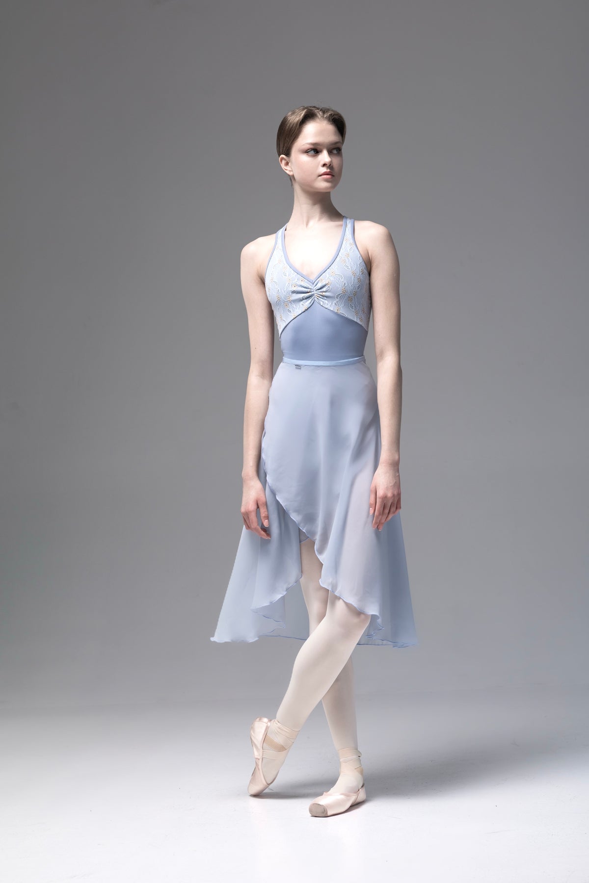 A dancer models a light blue sleeveless ballet dress with a sheer lace halter top, matching skirt, and white tights against a neutral gray studio background.