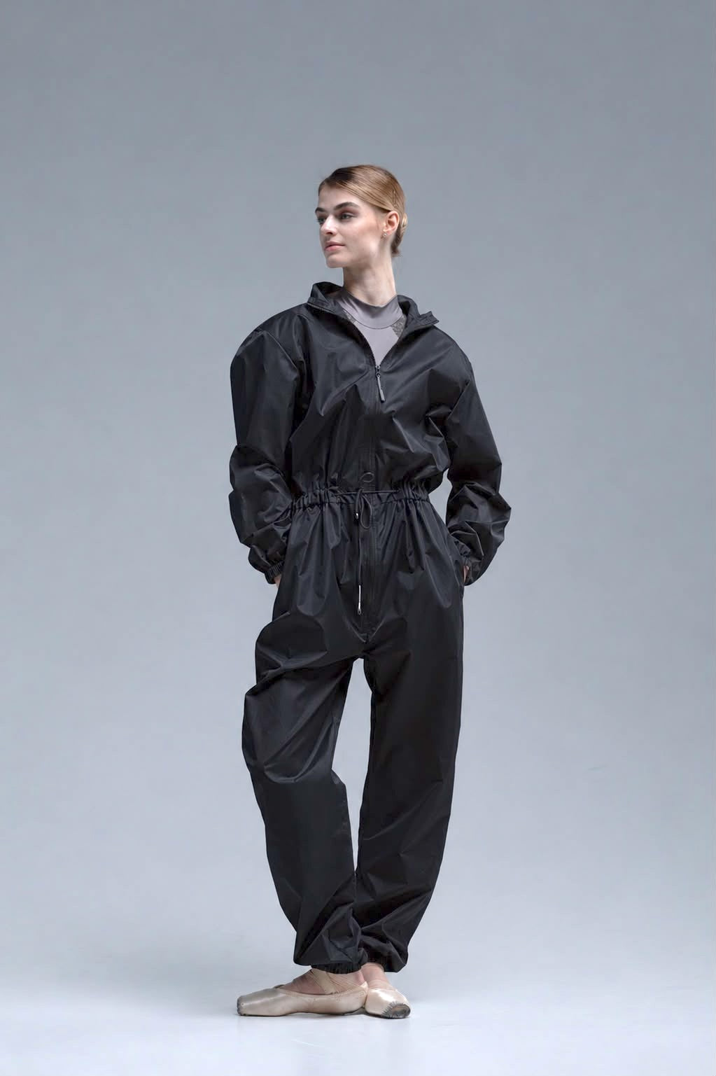 A ballerina wearing a black jumpsuit with a drawstring waist, styled over a gray leotard and beige ballet flats, standing against a neutral gray studio background.