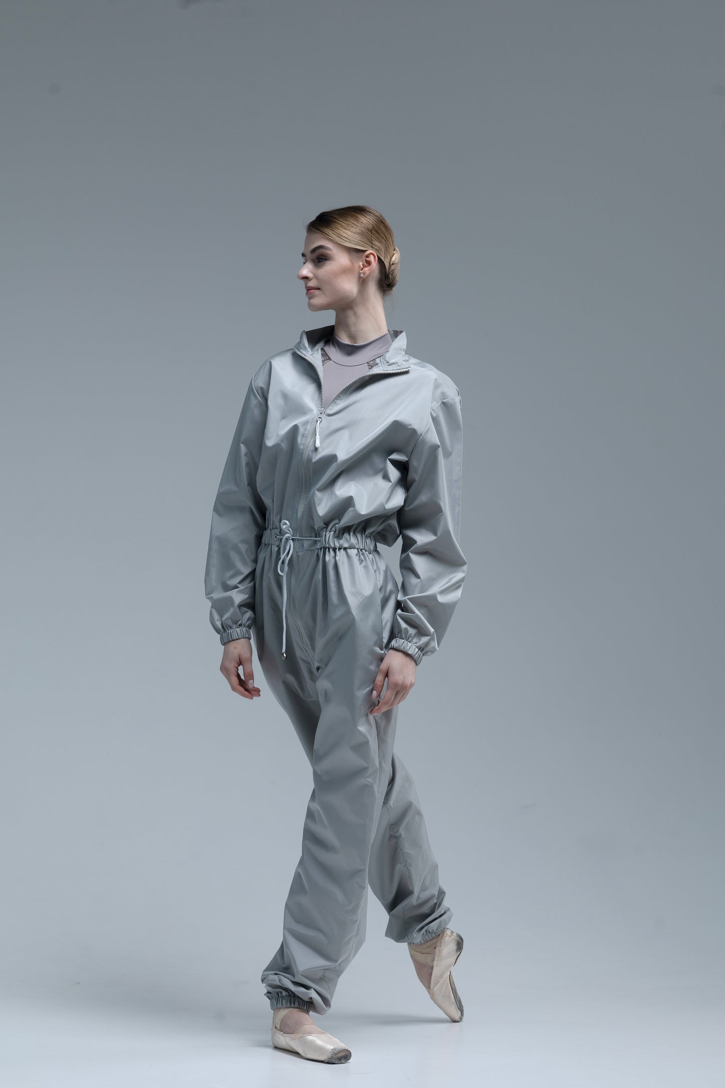 A model ballerina wearing a shiny light gray jumpsuit with a drawstring waist, standing in a studio against a solid gray background.