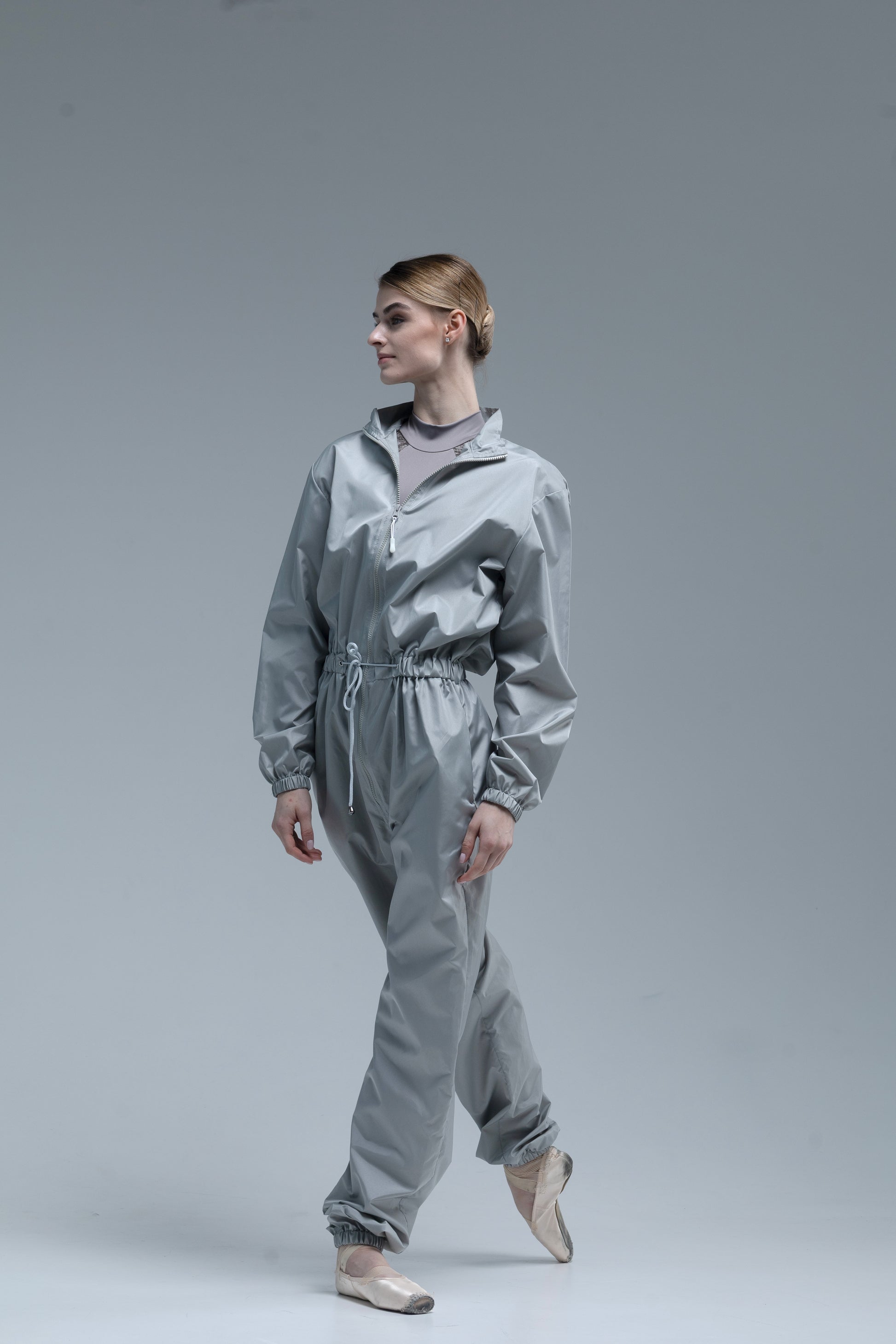 A model ballerina wearing a shiny light gray jumpsuit with a drawstring waist, standing in a studio against a solid gray background.