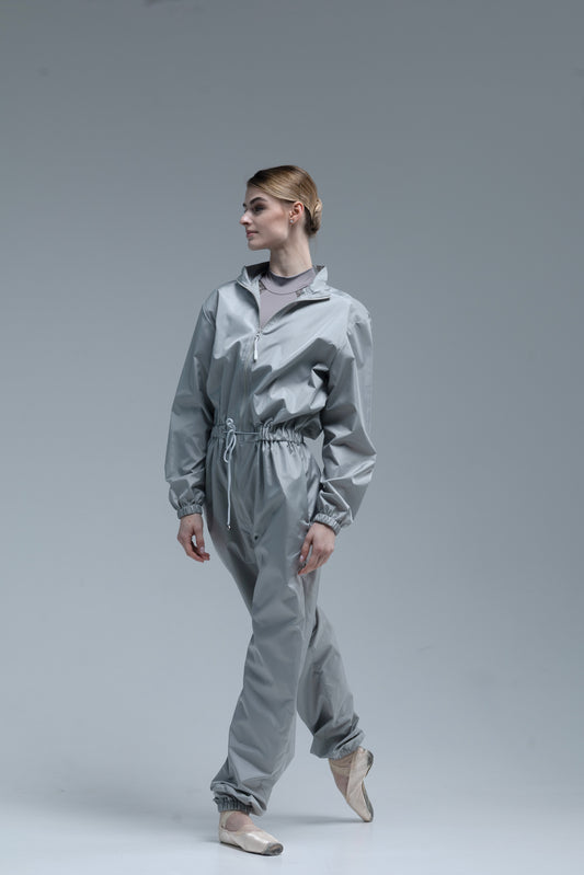 A model ballerina wearing a shiny light gray jumpsuit with a drawstring waist, standing in a studio against a solid gray background.