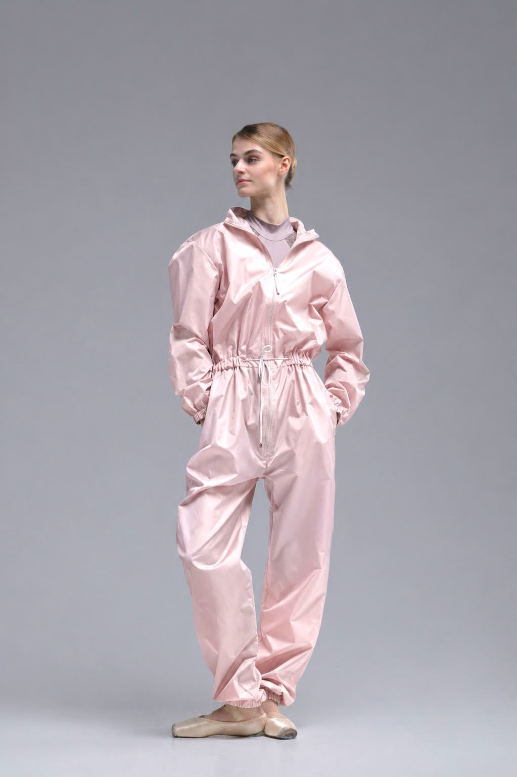 A ballerina wearing a shiny light pink one-piece jumpsuit with a hood and elastic waist, standing against a solid gray studio background.