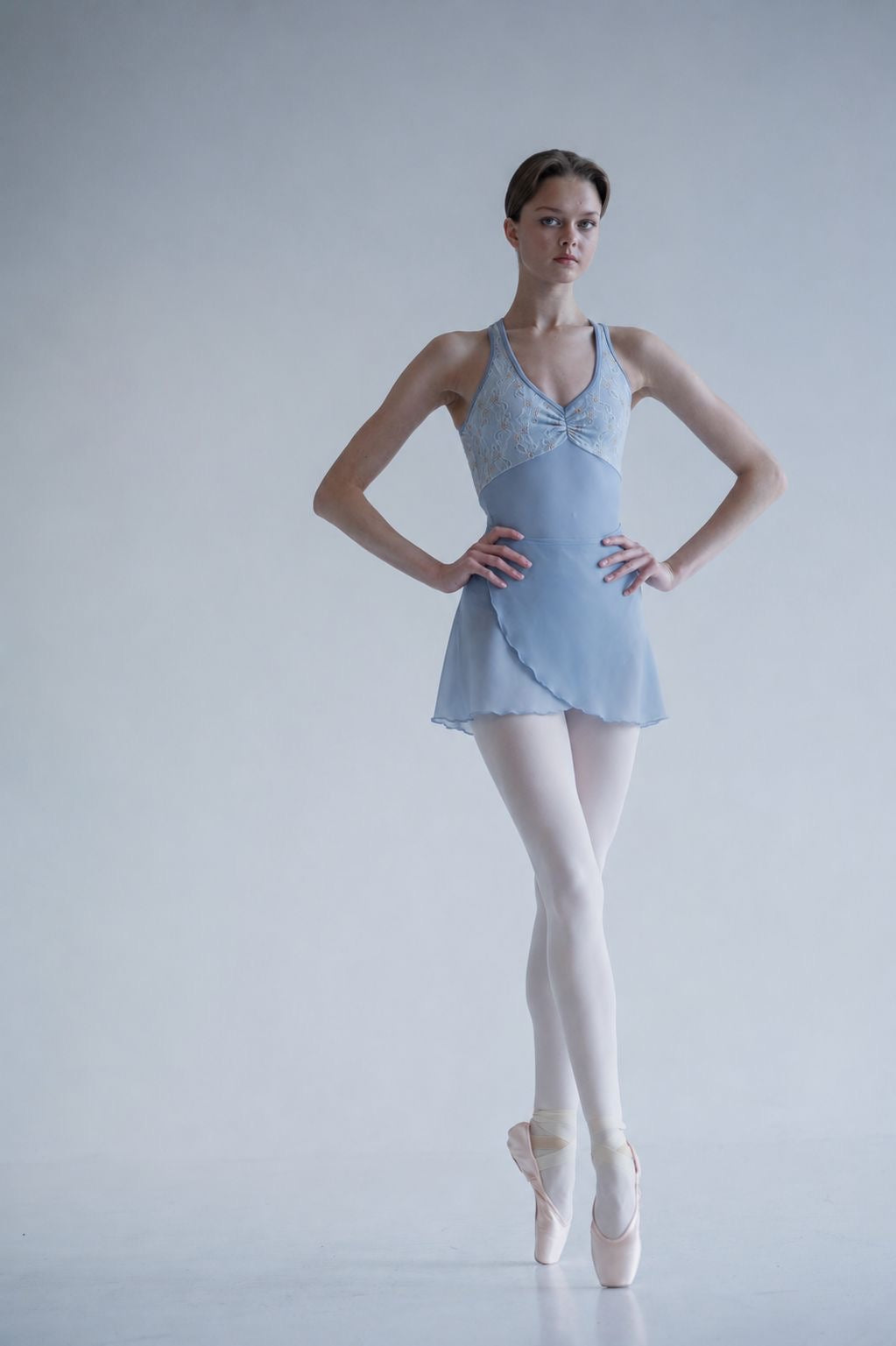 A female ballet dancer stands en pointe against a light gray studio background, wearing a pale blue sleeveless leotard, light blue skirt, white tights, and pink pointe shoes.