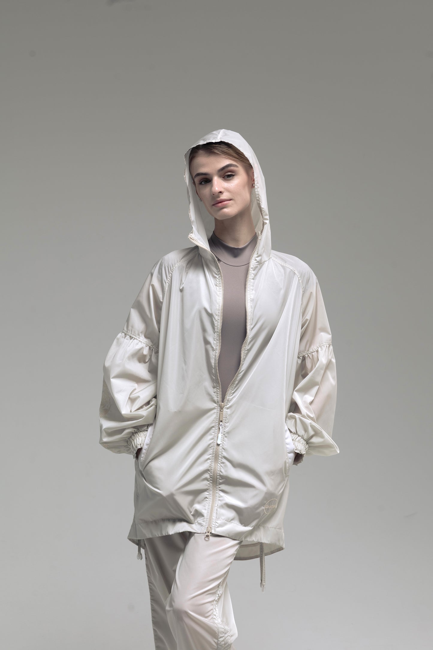 A ballerina wearing Embroidered suit ivory color, light beige hooded jacket with a front zipper, styled over a taupe top and matching light pants, set against a plain gray studio background.