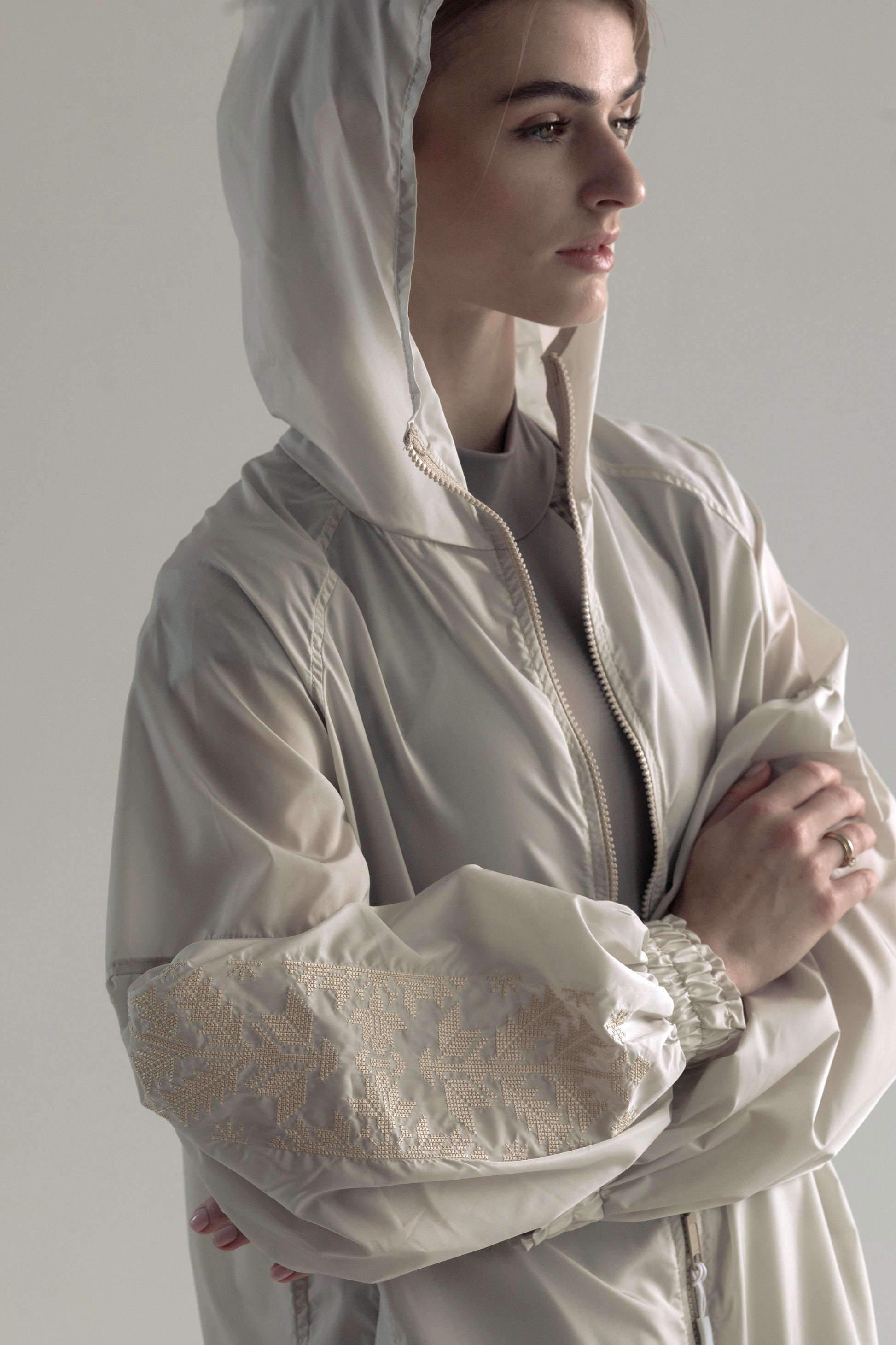 A ballerina wearing a light beige hooded jacket with intricate lace detailing on the sleeves, posed against a neutral light gray background.