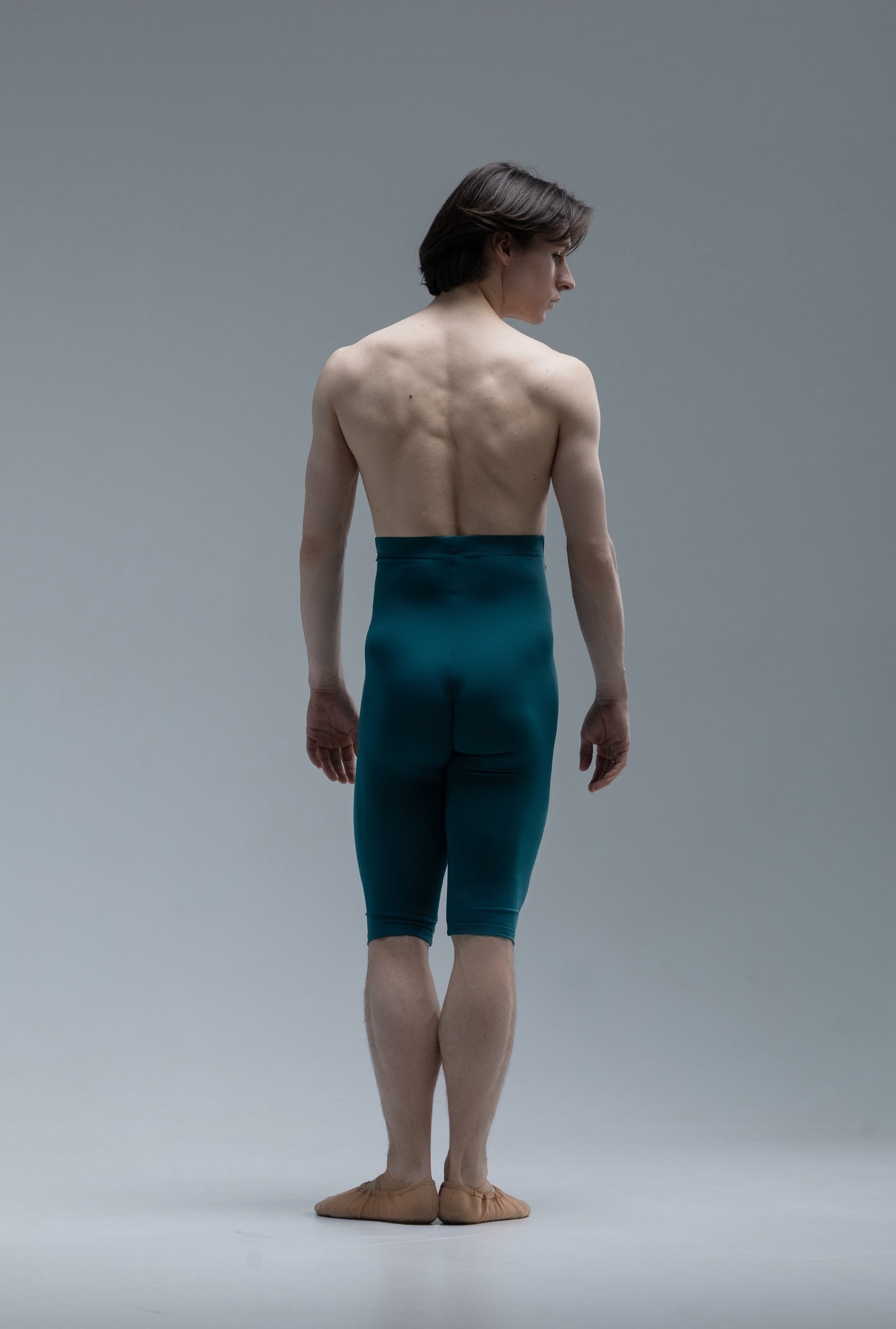 Ballet dancer in short teal tights and beige shoes back side on a gray background